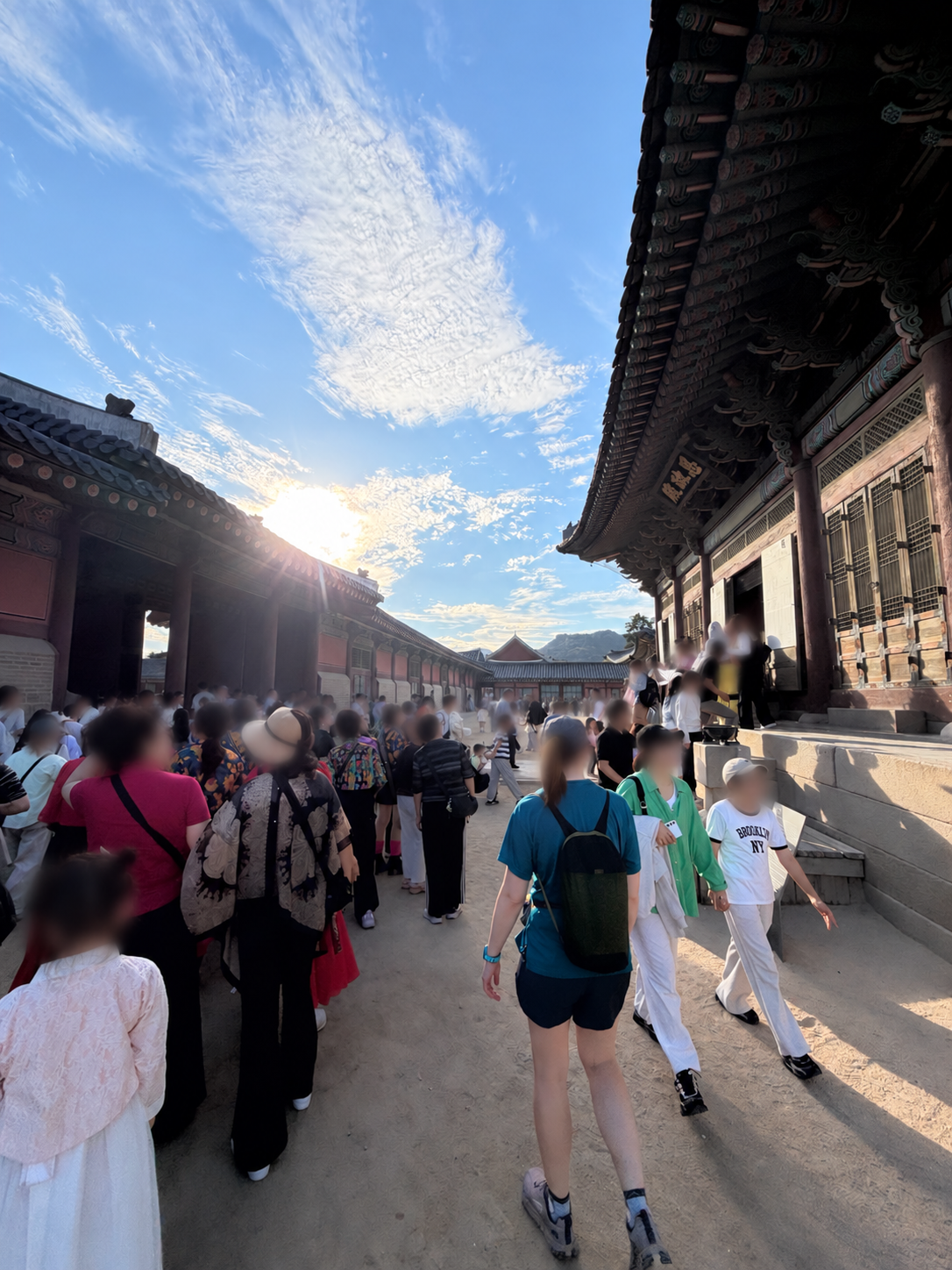 Visitors walking beside wooden palace halls at Gyeongbokgung beneath a bright sky; faces obscured for privacy